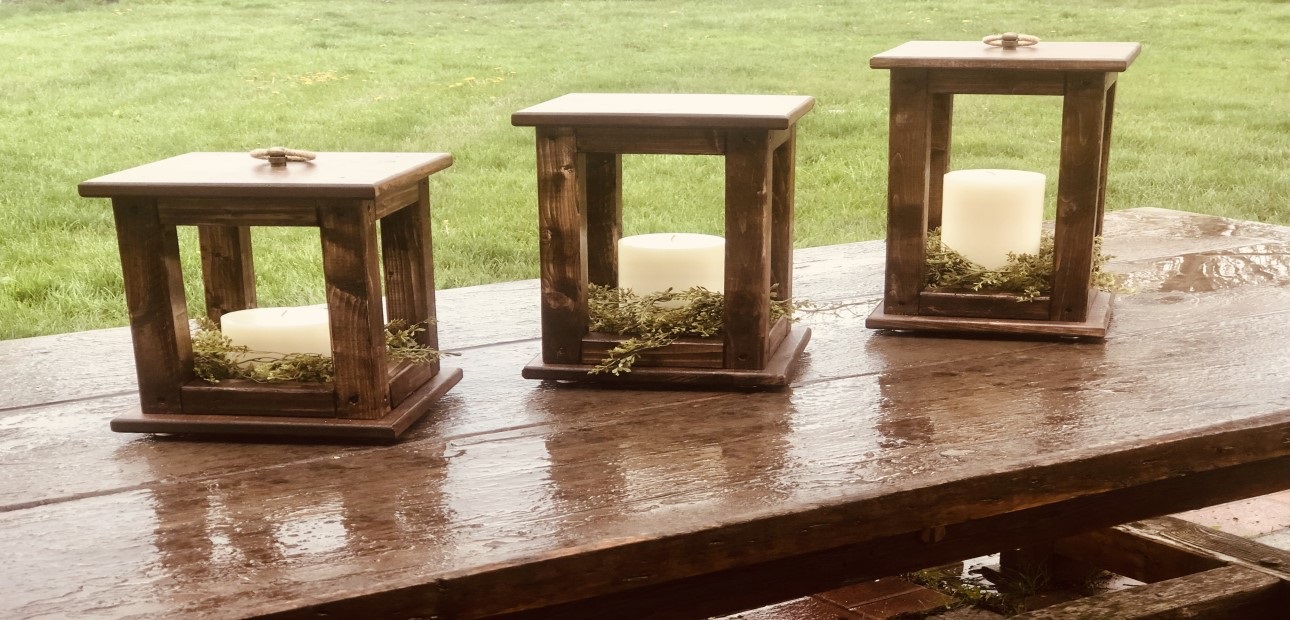 Three wooden lanterns holding candles on top of an outdoor wooden table.