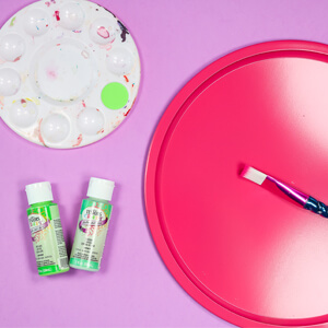 Dark pink pizza pan with paint brush, paint palette and two bottles of paint.