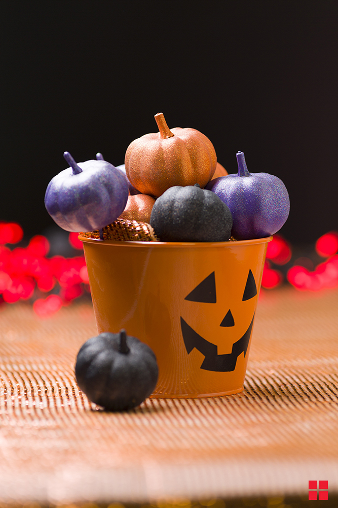 An orange metal pail with a jack-o-lantern face filled with decorative pumpkins painted in glittery orange, purple and black.