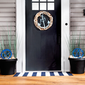 A nautical-themed front porch with an anchor wreath on the front door, urn planters filled with blue orbs and tall green grass and two wooden oars. 