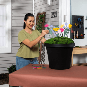 A woman placing painted wooden flowers in a black urn planter filled with green moss. 