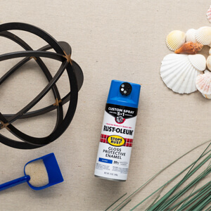 An unpainted metal orb, blue plastic sand shovel, pile of small seashells, green grass and a can of blue Rust-Oleum Stops Rust with Custom 5-in-1 spray paint. 
