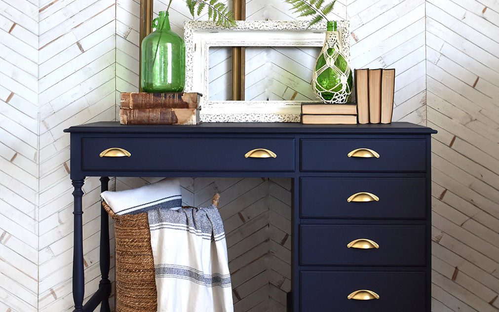 Navy blue desk with gold drawer handles, and books, vases and frames on top. 