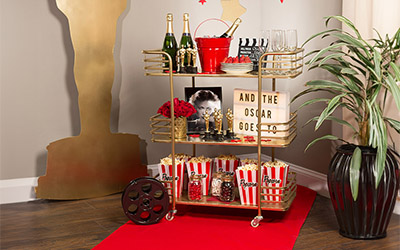 The corner of a room with a red carpet leading to a giant gold cut-out shaped like an Oscar award, and a golf bar cart full of champagne bottles, classes, flower and movie theater boxes of popcorn.