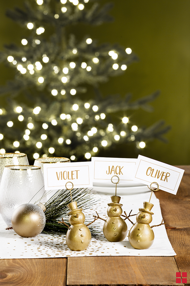 Three gold snowman place card holders with cards reading Violet, Jack and Oliver on a table with a Christmas tree in the background.