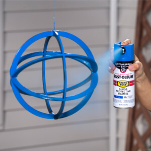 Close-up of a hand applying blue Rust-Oleum Stops Rust Custom Spray 5-in-1 spray paint to a metal orb.