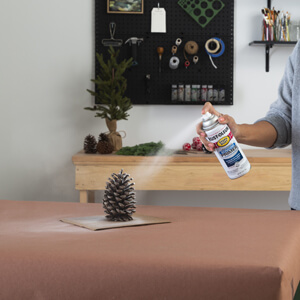 A woman applying spray paint primer to a pine cone.