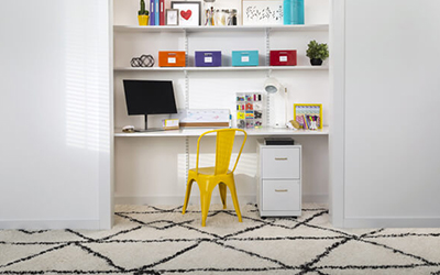 A yellow chair and white shelves holding a computer, storage boxes and office supplies inside a closet without doors. 