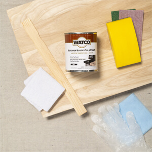 A can of Watco Butcher Block Oil + Stain, stir stick, sand paper and other supplies on an unfinished wood board.