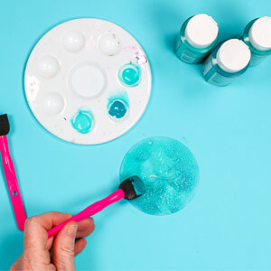 A hand using a brush to apply blue paint to a flat, circular ornament. 