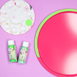 Dark pink pizza pan with painters tape around ends, paint palette and a bottle of paint.