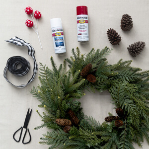 A green wreath with pine cones, two cans of Rust-Oleum spray paint, a spool of ribbon, decorative picks and a pair of scissors. 