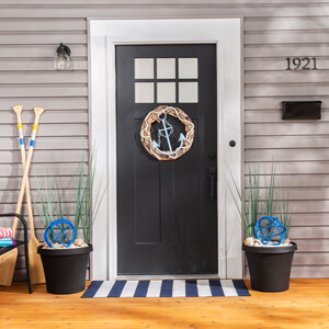 A nautical-themed front porch with an anchor wreath on the front door, urn planters filled with blue orbs and tall green grass on either side of the door and two wooden oars to the left of the door. Striped beach towels and a beach bag sit on a bench on the left side of the porch. 