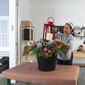  A woman placing a red lantern with a candle on top of a black planter filled with greenery and pine cones. 