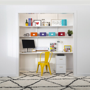 A yellow chair and white shelves holding a computer, storage boxes and office supplies inside a closet without doors.