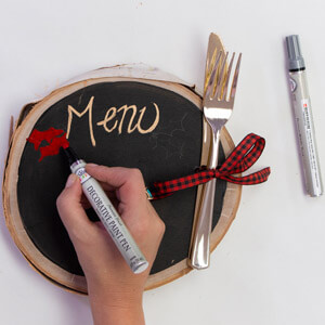 A hand using a Testors paint pen to write on a chalkboard menu.