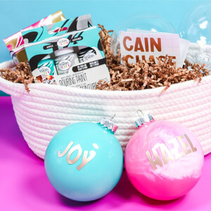 A blue ornament that says “Joy” and a pink ornament that says “Hazel” in front of a white basket containing Testors paints, glass ornaments and letter stickers. 